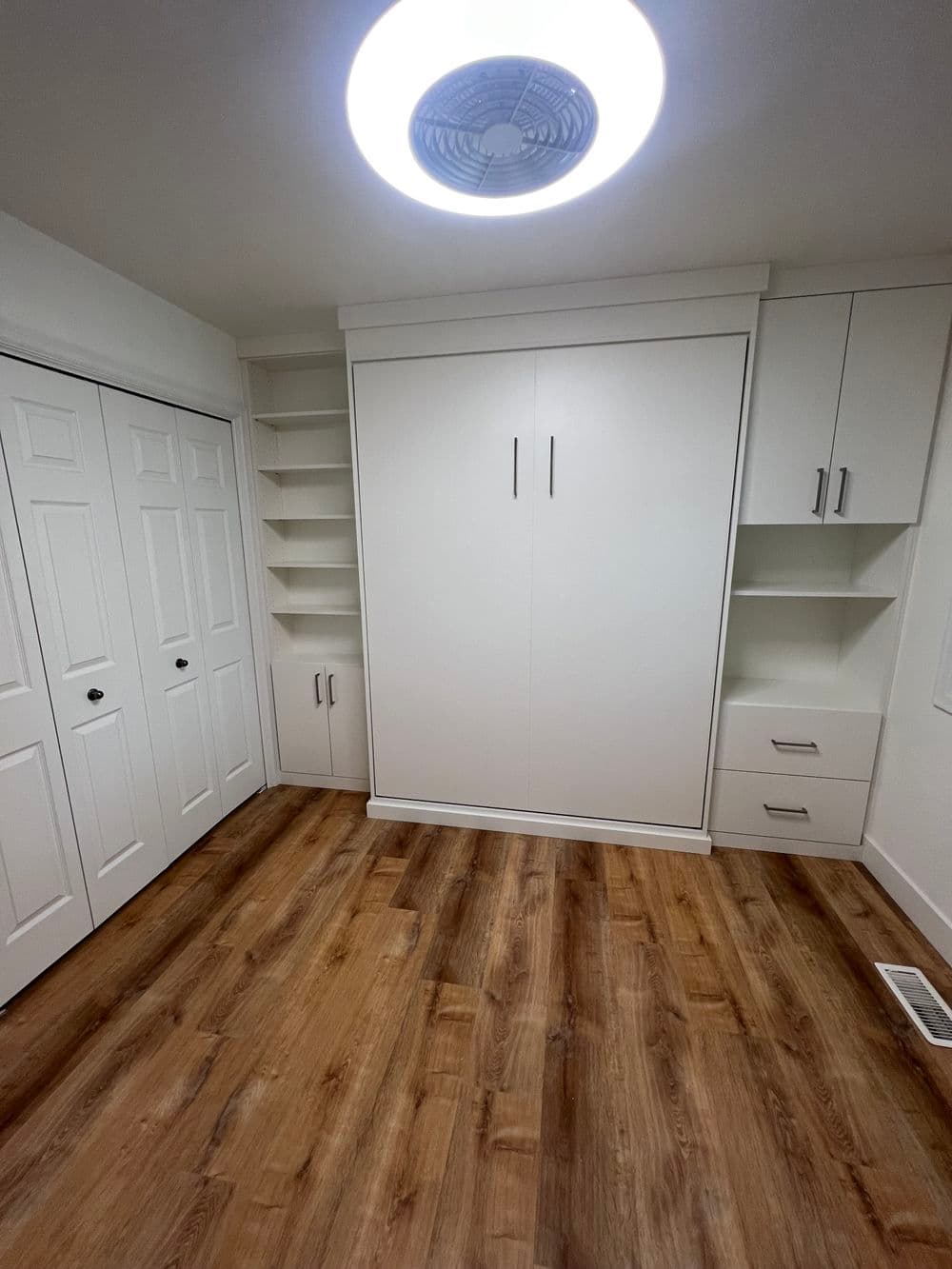 Modern bedroom with a built-in wardrobe, shelving, and wooden flooring illuminated by a ceiling light.