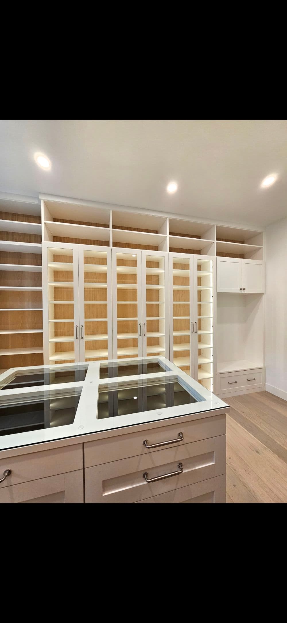 Modern walk-in closet with white shelves, glass display, and warm wood accents. Spacious design.